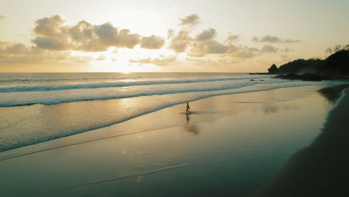 Persona caminando por una amplia playa de arena al atardecer, con olas suaves y un promontorio rocoso