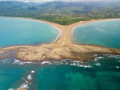 Vista aérea de un tómbolo de arena con forma de cola de ballena entre aguas turquesa, con colinas boscosas al fondo