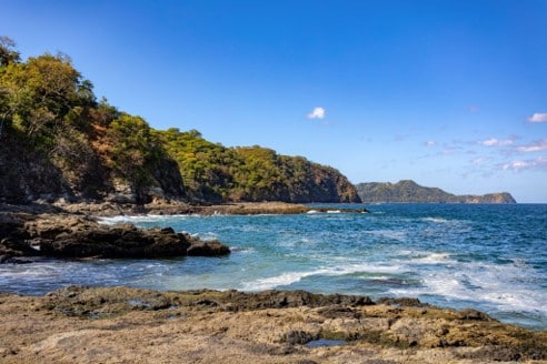 Costa rocosa con oleaje y acantilados boscosos bajo un cielo azul despejado