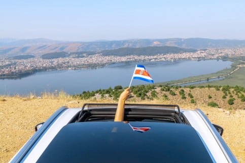 Mano sosteniendo una bandera de Costa Rica desde el techo corredizo de un carro con vista a un lago y un pueblo