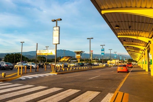 Zona de llegadas de un aeropuerto con techo amarillo, carros estacionados y montañas a lo lejos