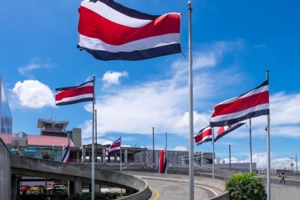 Banderas de Costa Rica ondeando en astas junto a la terminal de un aeropuerto bajo un cielo azul